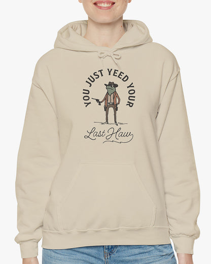 Woman wearing sand beige 'You Just Yeed Your Last Haw' hoodie with funny retro cowboy frog graphic