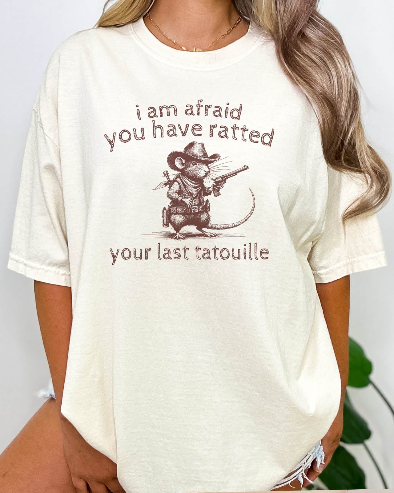 Woman wearing "You Have Ratted Your Last Tatouille" Shirt in natural beige color with a retro cowboy rat graphic