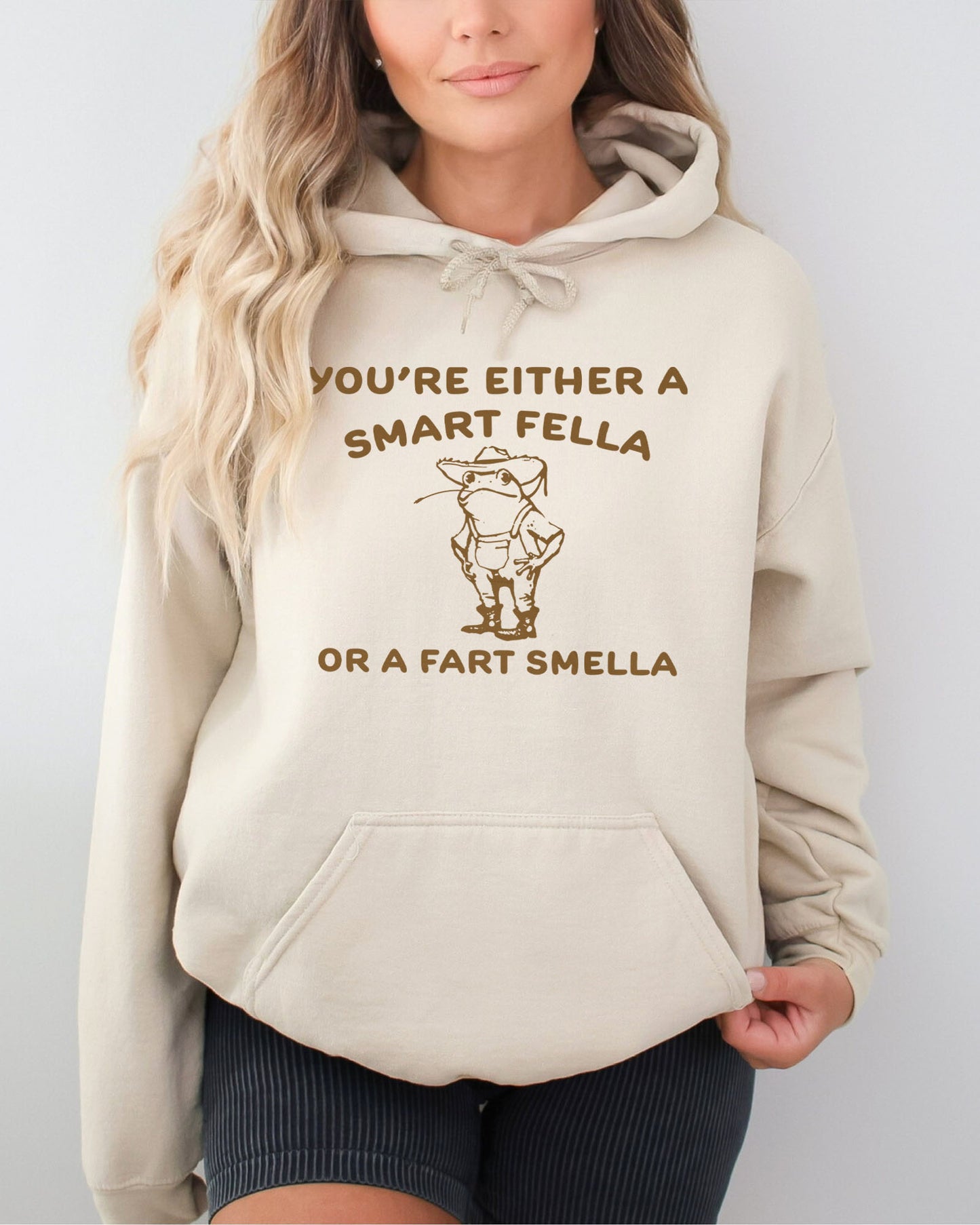 Woman wearing a sand beige hoodie featuring a funny retro-style country frog graphic with the quote 'You're Either A Smart Fella Or A Fart Smella'.