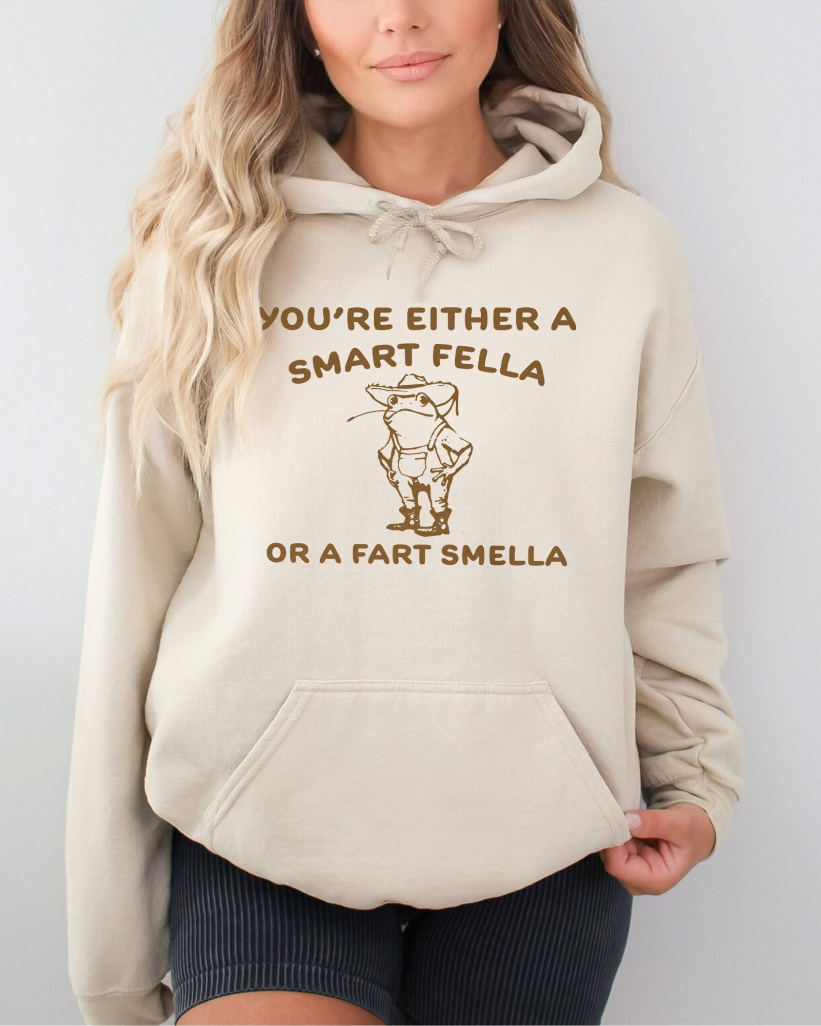 Woman wearing a sand beige hoodie featuring a funny retro-style country frog graphic with the quote 'You're Either A Smart Fella Or A Fart Smella'.