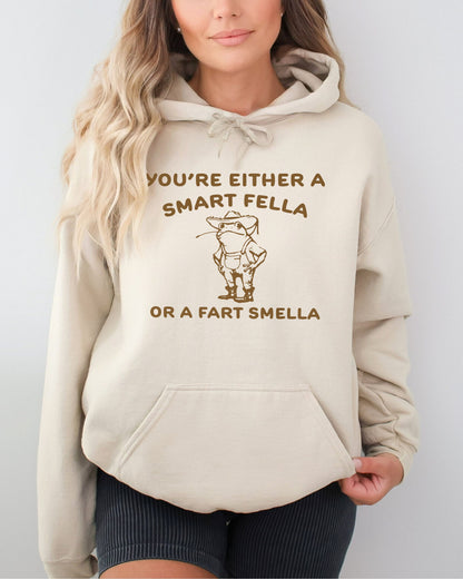Woman wearing a sand beige hoodie featuring a funny retro-style country frog graphic with the quote 'You're Either A Smart Fella Or A Fart Smella'.
