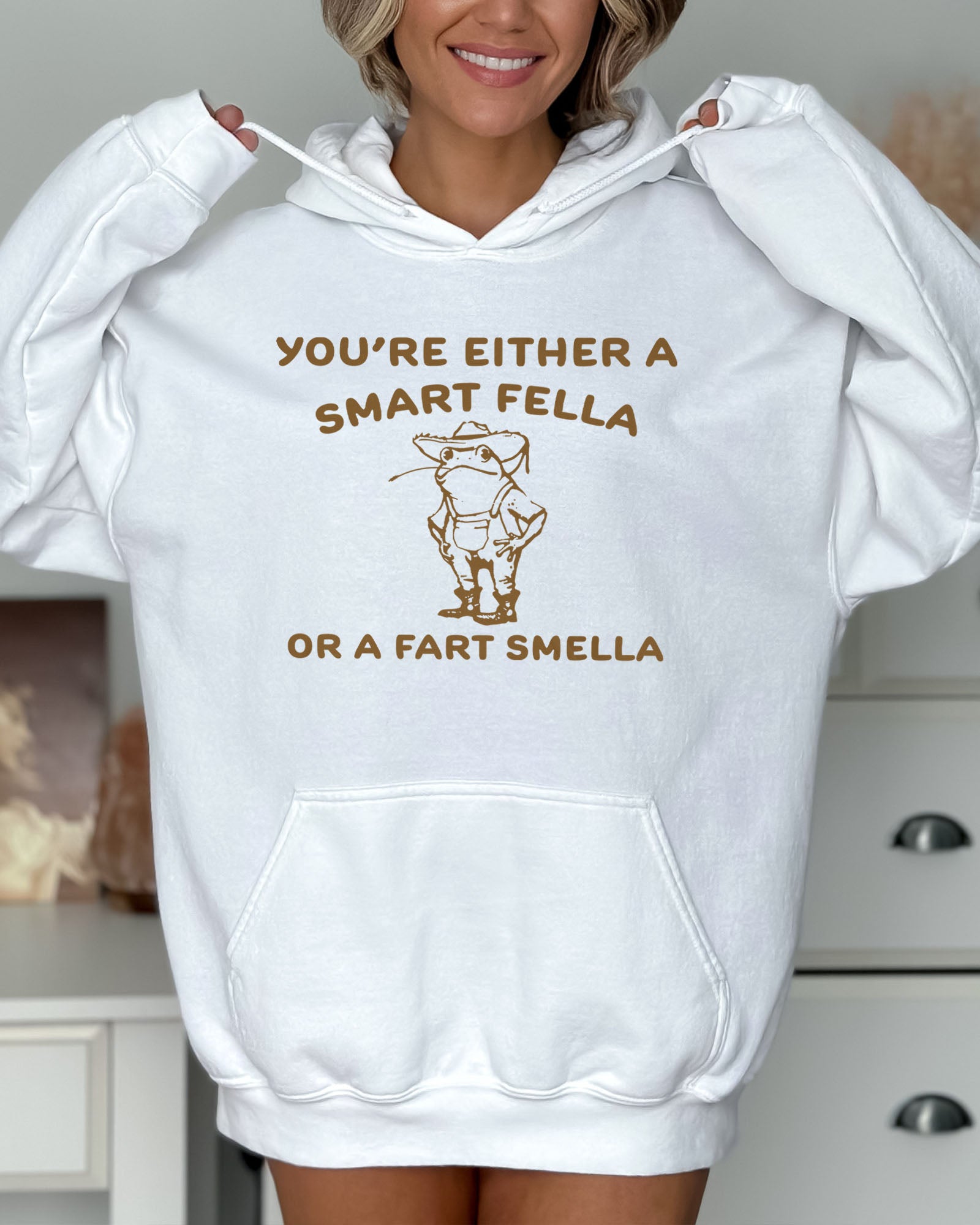 Woman wearing a white hoodie featuring a funny retro-style country frog graphic with the quote 'You're Either A Smart Fella Or A Fart Smella'.