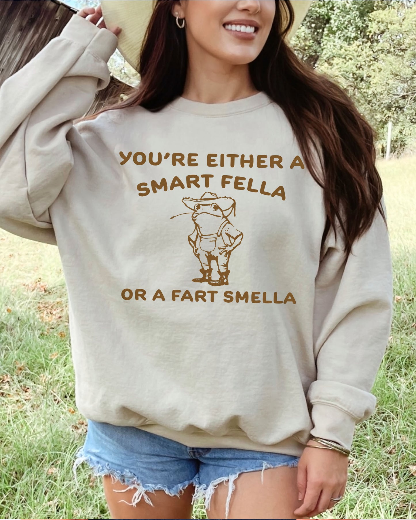 Woman wearing a sand beige sweatshirt featuring a funny retro country frog graphic and the quote 'You're Either A Smart Fella Or A Fart Smella'.