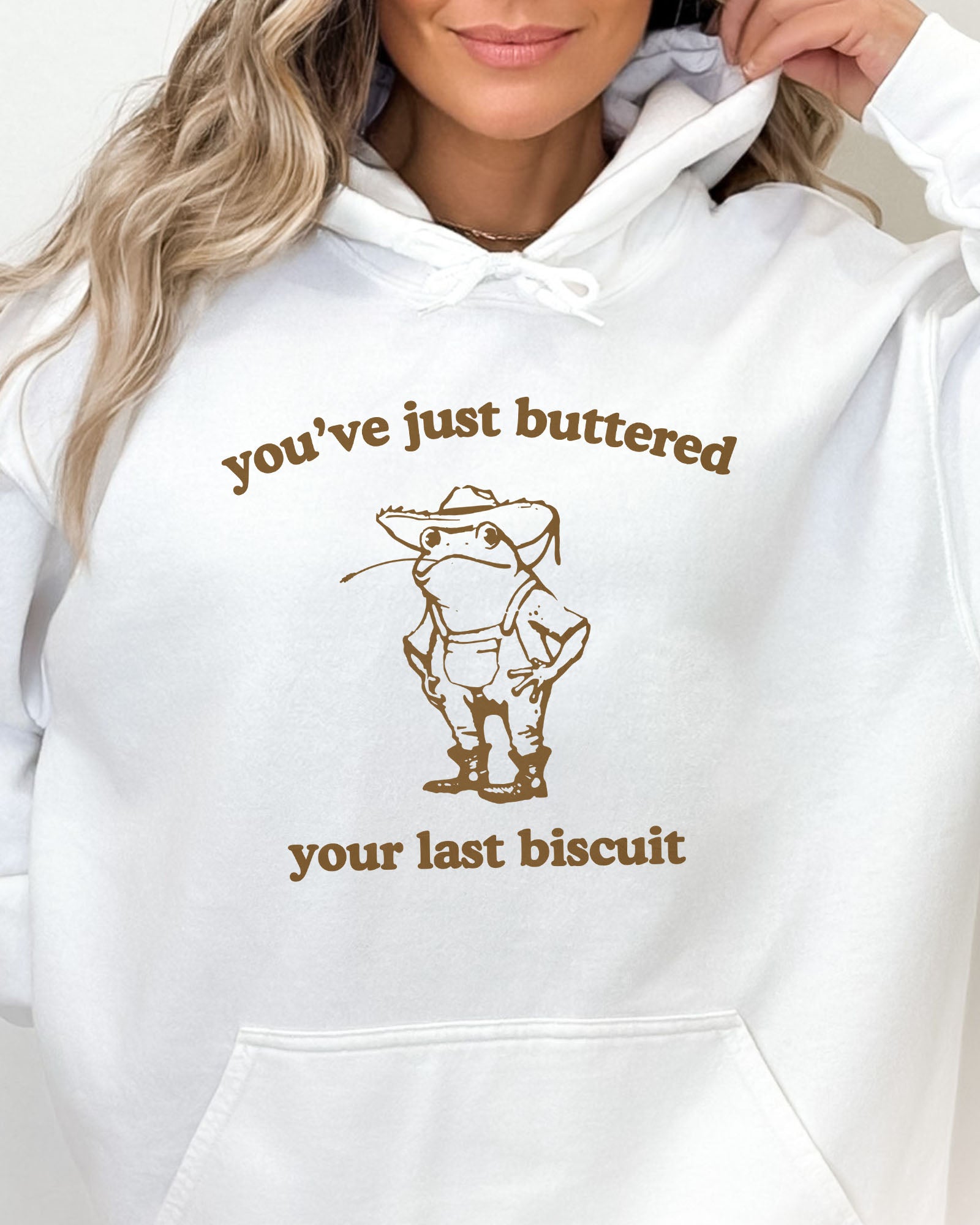 Close-up of a white hoodie featuring a vintage country frog graphic with the quote 'You've Just Buttered Your Last Biscuit'.