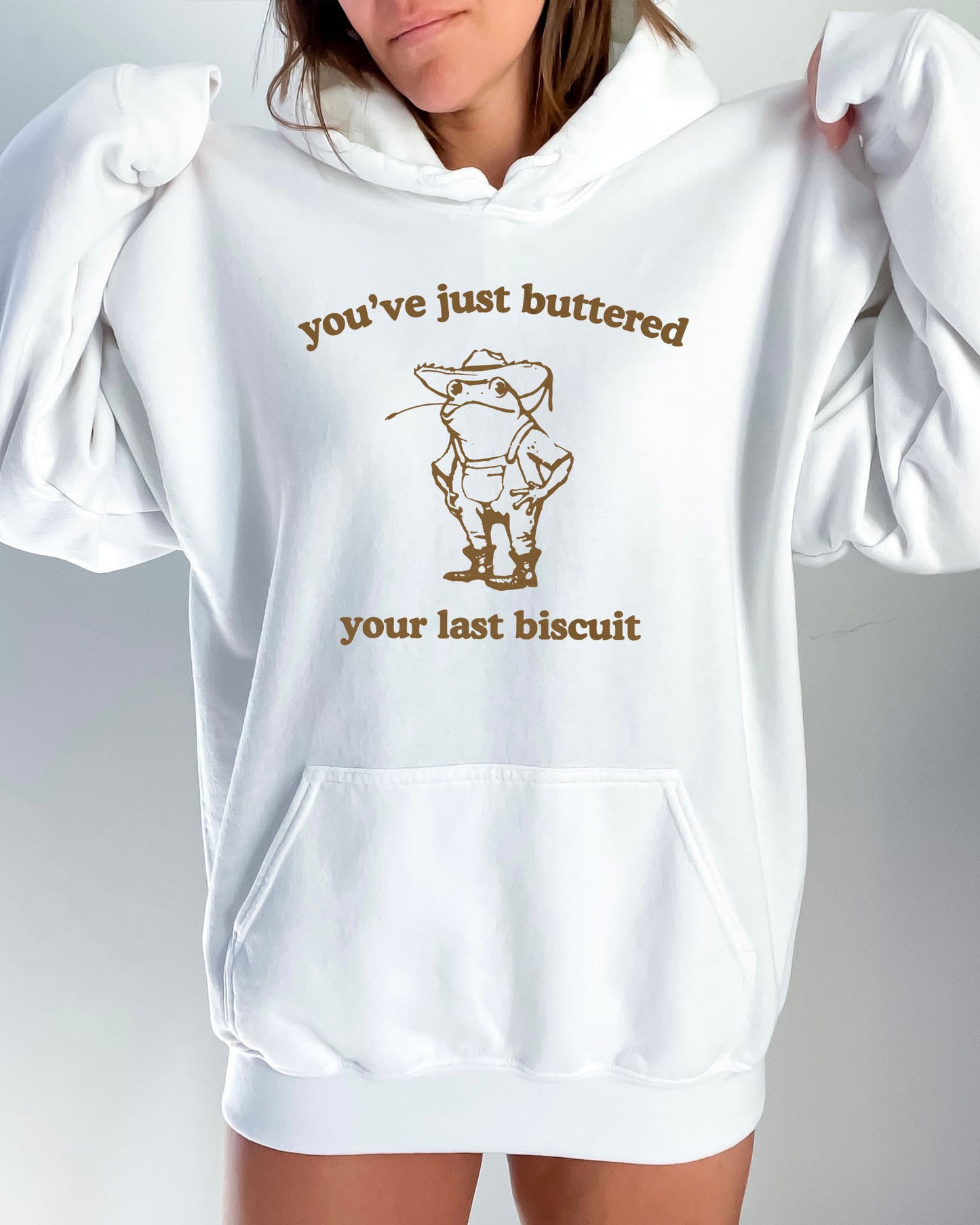 Woman in a white hoodie featuring a vintage country frog graphic with the quote 'You've Just Buttered Your Last Biscuit'.