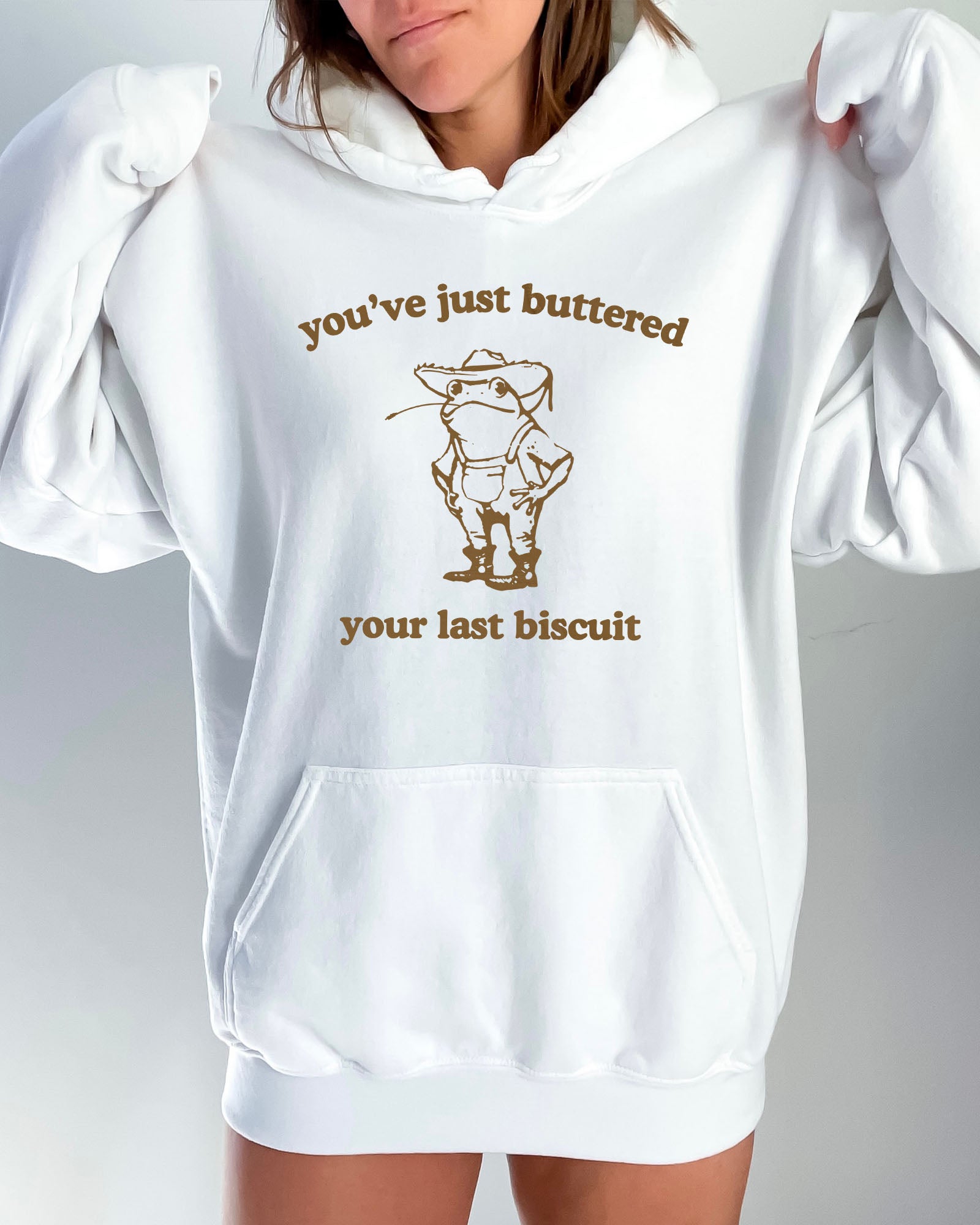 Woman in a white hoodie featuring a vintage country frog graphic with the quote 'You've Just Buttered Your Last Biscuit'.